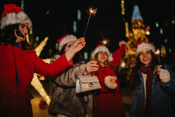 Fototapeta premium Four girls wearing Santa hats hold lit sparklers and smile at night. Friends celebrate outdoors amid festive lights and holiday decorations.