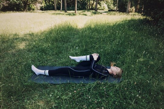 Woman in athletic wear lies on mat in tall green grass pulling knee to chest for flexibility. Post workout stretch, recovery stretch outdoor, cool down routine grass, Muscle recovery position.