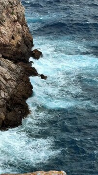 Dramatic vertical view of waves crashing against rocky shoreline under gray storm clouds near Capdepera, Mallorca. Concept of nature power, seascape, rugged coast, travel. 