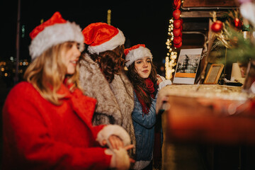 Fototapeta premium Three girls wearing Santa hats browse a festive outdoor Christmas market stall. They look at holiday treats and decorations under warm market lights.