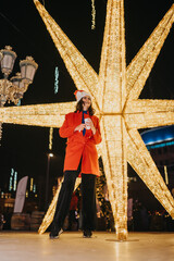 Fototapeta premium Young woman in a Santa hat and red coat holds a takeaway cup in front of a giant illuminated star. Festive night scene shows her smiling and enjoying holiday lights at an outdoor market.