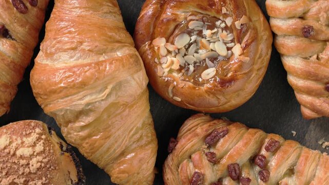 Top view closeup of assortment of freshly baked sweet pastries and buttery croissants, caramel bun, slowly rotating on dark wooden background