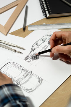 Hands of engineer sketching automotive vehicle prototype with pencil on paper at desk. Industrial designer developing new car blueprint drafts near measuring tools and rulers.