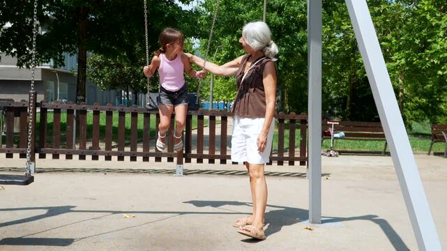 Loving grandmother gently pushing her happy granddaughter on a swing at the park, enjoying a beautiful sunny day together and creating precious family memories during a summer afternoon