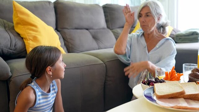 Affectionate grandmother and her young granddaughter spending quality time together, playing, talking, and enjoying a snack in the living room, strengthening their intergenerational family bond