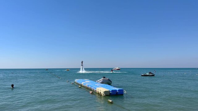 Turkey. 21.07.2026 Man riding flyboard above sea water. Athlete performing water sport stunt in ocean. Summer vacation activity with jet ski boat and floating dock at blue sky.