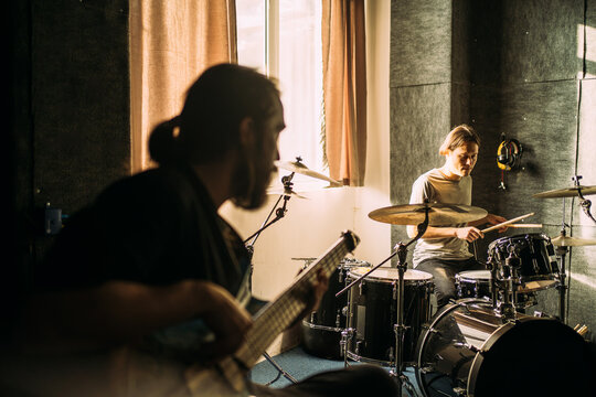 Drummer rehearsing in music studio while bassist plays in foreground, live band practice, drum kit performance, recording session, creative collaboration.