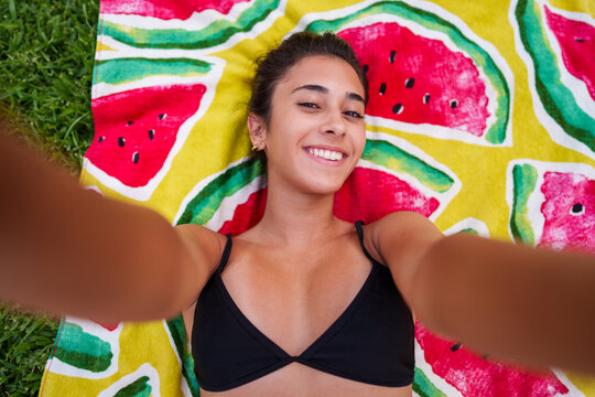 Young woman taking a happy selfie while lying on a vibrant watermelon print towel enjoying summer vacation