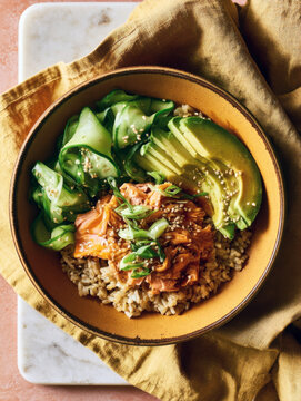 Healthy poke bowl dish featuring cooked salmon fillet, sliced avocado, ribboned cucumber, brown rice, green onions, and sesame seeds served in a ceramic bowl over an orange napkin on marble backdrop