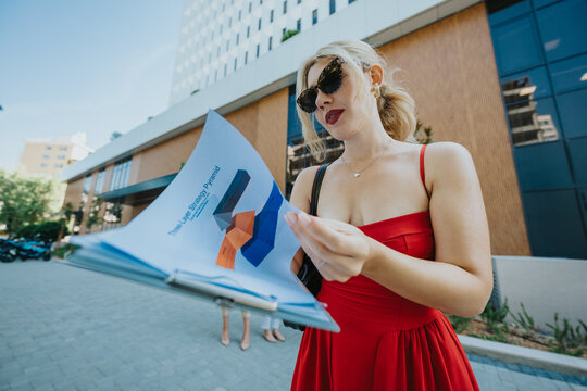 Young woman in a red dress reads a printed business report while standing outdoors. She flips through documents in front of a modern office building wearing sunglasses.