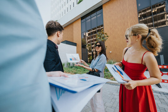 Young professionals review charts and reports during an outdoor business meeting. A small team discusses a presentation outside a modern office building.