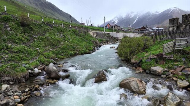 Fast flowing mountain river in Ushguli Svaneti Georgia with snowy peaks, rocky banks, village houses, turbulent water current, travel