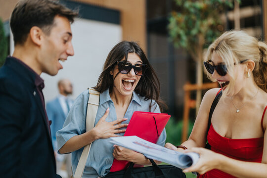 Three young friends laugh and react excitedly as they look at papers and a red folder. A surprised woman holds the folder while her friends share the joyful moment outdoors.
