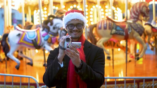 Smiling man wearing a santa hat filming with a retro camcorder at a christmas funfair with a carousel in the background