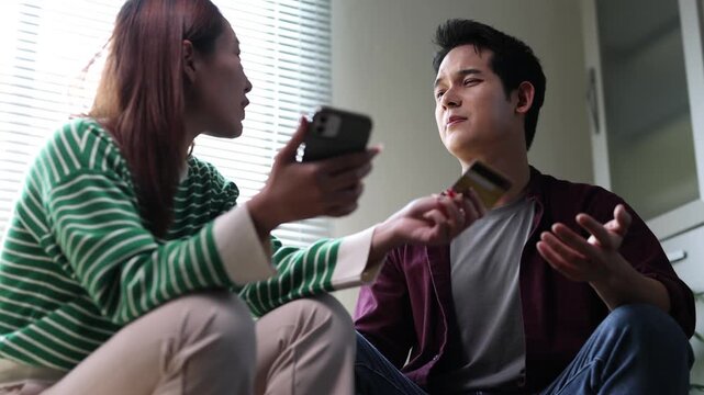 Worried asian couple arguing about money holding smartphones and discussing home finance problems while sitting on a sofa in the living room at home.
