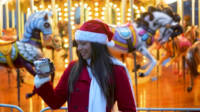 Young woman wearing a santa hat filming a vintage carousel with a retro camcorder at a christmas fair