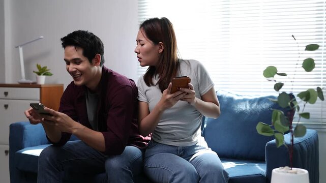 Jealous girlfriend secretly looks at boyfriend's smartphone screen while they sit together on the living room sofa.