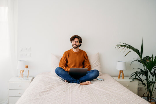 Happy freelancer with laptop sitting on bed at home