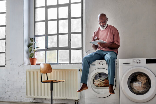 Bald man using tablet PC sitting on washing machine at home