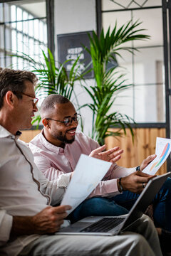 Two businessmen using laptop and discussing documents on sofa in loft office