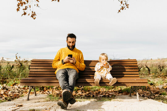 Father and daughter sitting on a bench in the park in autumn, father using smartphone, daughter eating an apple
