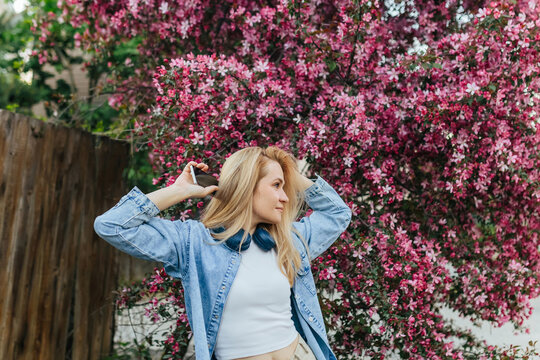 Blonde woman holds smartphone near blooming pink tree outside