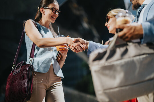 Two business professionals shake hands outdoors while holding coffee and brief bags. The friendly negotiation is captured in a candid networking moment.