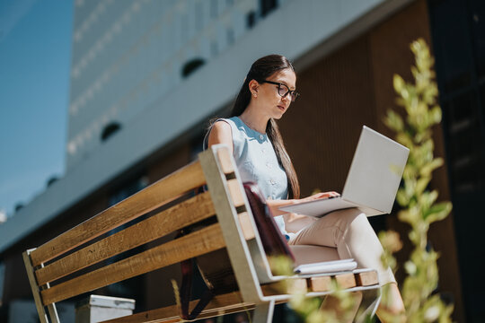 A young woman sits on a bench using a laptop, concentrating on her work. She works outdoors in an urban setting on a sunny day, typing on her computer.