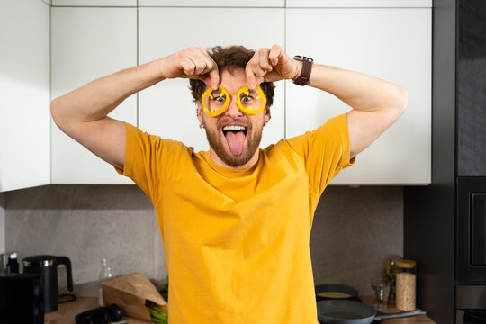 Playful man sticking out tongue while holding bell pepper slices on eyes in kitchen