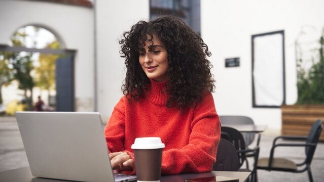 Beautiful Caucasian young woman with curly hair working on laptop outdoors at cafe. Female freelancer sitting at wooden table with coffee cup and flowers, smiling while using computer outside.