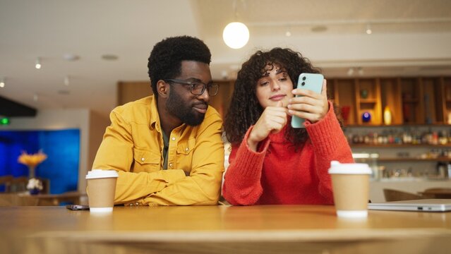 Mixed-races man in yellow shirt and woman in red sweater sitting at cafe table with coffee cups, looking at smartphone together. Male with glasses watching attentively as female shows phone screen.