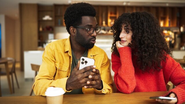 Man in yellow shirt showing smartphone to woman in red sweater at cafe table. African American male with glasses holding phone while curly-haired female listens with interest, both sitting with coffee