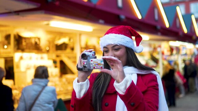 Cheerful woman wearing a santa hat filming with a retro camcorder at a festive outdoor christmas market with glowing lights