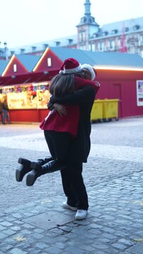 Romantic couple wearing santa hats hugging and spinning at christmas market, enjoying their winter holiday vacation together