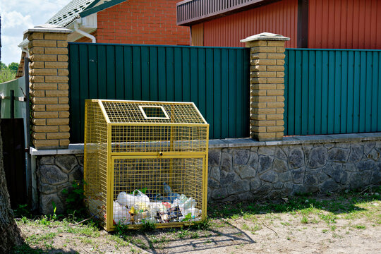 A yellow wire mesh waste container for sorting plastic bottles installed near a fence in a residential area.