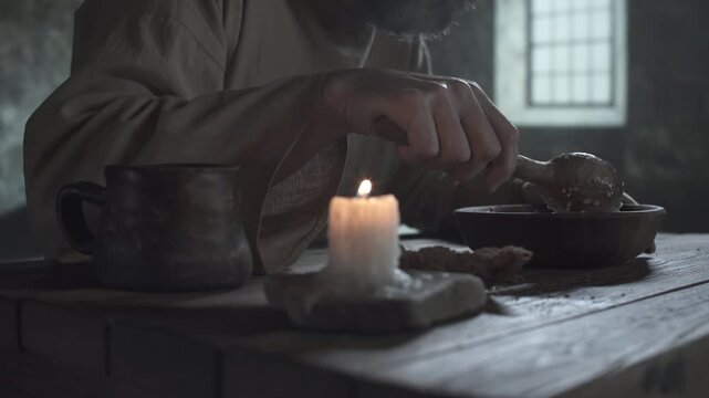 Close up of the hands of a medieval prisoner scooping porridge with a wooden spoon from a bowl inside a gloomy cell, with a blurred candle and a clay mug in the foreground