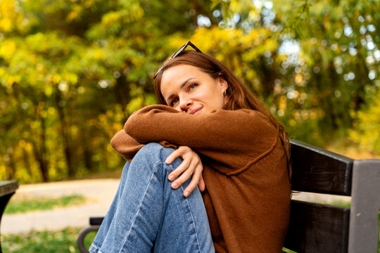 Woman in brown sweater embracing herself while enjoying cozy moment alone on bench in peaceful autumn park.