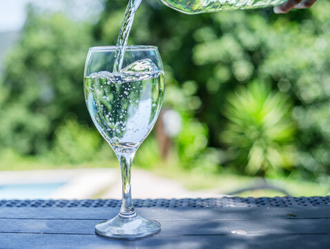Pouring clear water into a wine glass near swimming pool outdoors in garden.