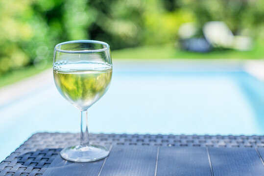 Glass of white wine on dark woven table near swimming pool outdoors