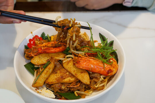 Chopsticks lift Char kway teow with shrimp and vegetables. Char kway teow is a stir-fried rice noodle dish from Maritime Southeast Asia. In Hokkien and Teochew, char means stir-fried and kway teow ref