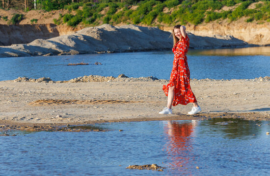 Young woman in red summer dress walking by blue lake on a sunny day