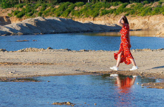 Young woman in red summer dress walking by blue lake on a sunny day