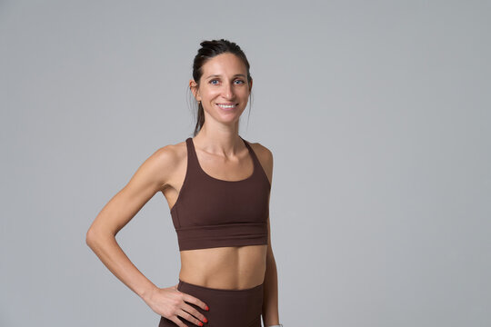 Smiling fit woman posing in brown yoga set standing against gray wall