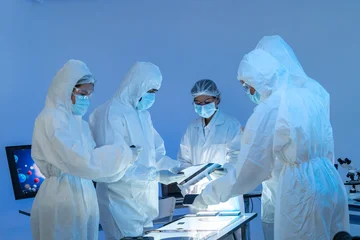 Four specialists in white protective suits and masks gathered around an inspection light table, semiconductor production teamwork in blue cleanroom. © Jestercine