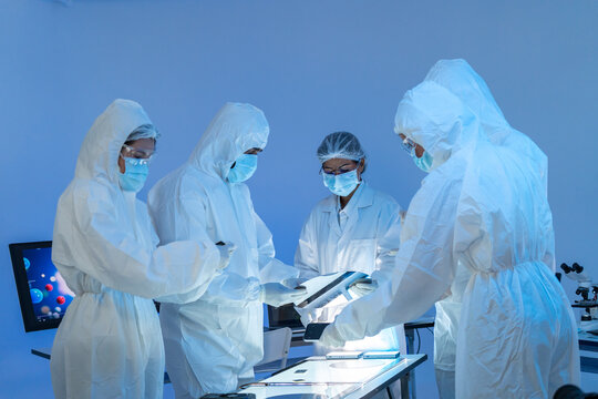 Four specialists in white protective suits and masks gathered around an inspection light table, semiconductor production teamwork in blue cleanroom.