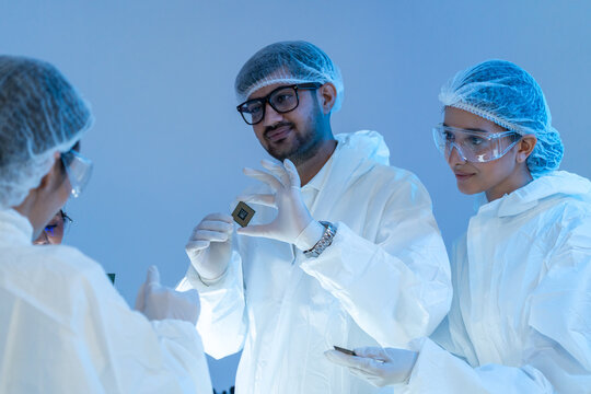Man in glasses and mask holding a small processor chip while two female colleagues in protective suits look on, semiconductor teamwork in cleanroom.