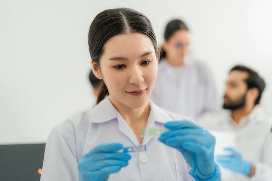 Young Asian woman in lab coat and gloves inspecting a green and blue specimen slide, research colleagues talking in background, focused lab work.