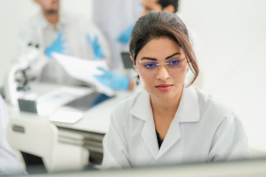 Young woman in eyeglasses and lab coat concentrating at a screen while colleagues review documents behind, science research in lab.