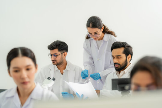 Five scientists in lab coats and gloves studying paperwork and samples around a microscope, collaborative team discussion in research lab.