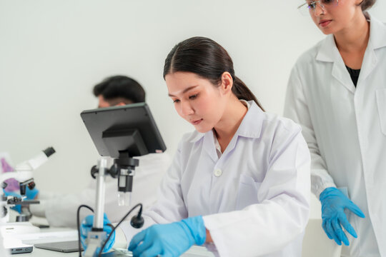 Young Asian woman in lab coat focused at a digital microscope while colleague observes, research teamwork at the bench in laboratory.